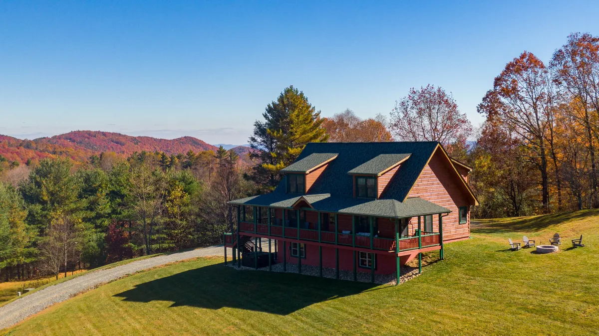 The image features a newly built log cabin-style house nestled in the mountains near Boone, North Carolina. The home is surrounded by vibrant autumn foliage, with sweeping long-range mountain views creating a stunning backdrop. In the foreground, a cozy fire pit area invites guests to gather outdoors, while the spacious porch is perfect for relaxing and soaking in the scenery.