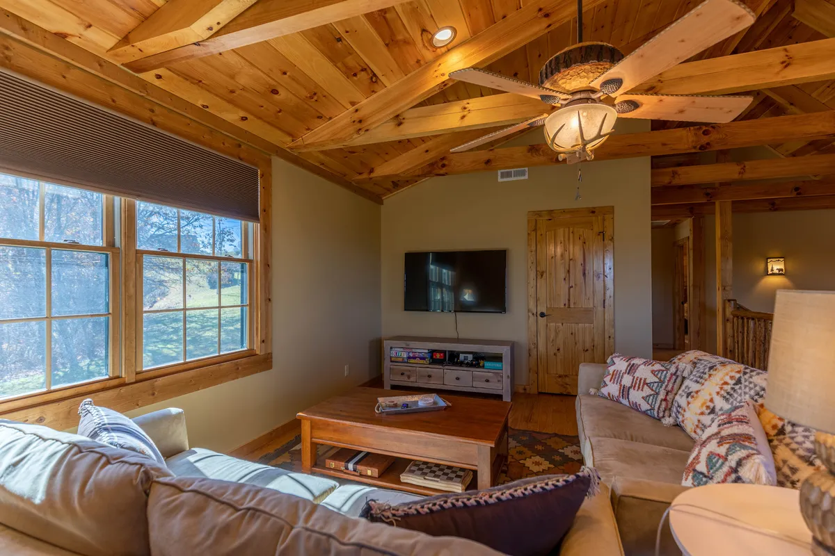 A bright, inviting living space with a wooden ceiling, large windows connecting to the outdoors, comfortable sofas around a coffee table, and a television. The layout encourages group relaxation, socializing, and enjoying both indoor comfort and outdoor views.