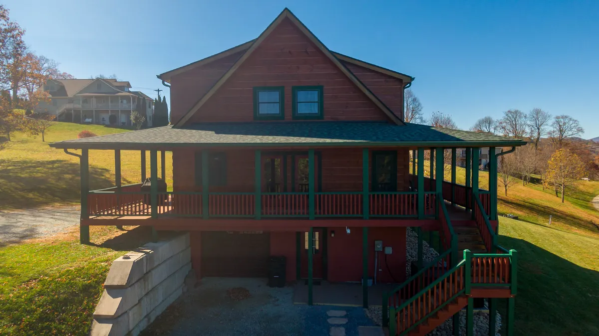 A wide view of the large front porch with wooden exterior and green trim, set against a backdrop of natural landscaping. The porch is a comfortable spot for relaxing, enjoying morning coffee, or watching kids and pets play in the yard. The setting emphasizes a seamless connection between the home and nature, suitable for families with multiple dogs.