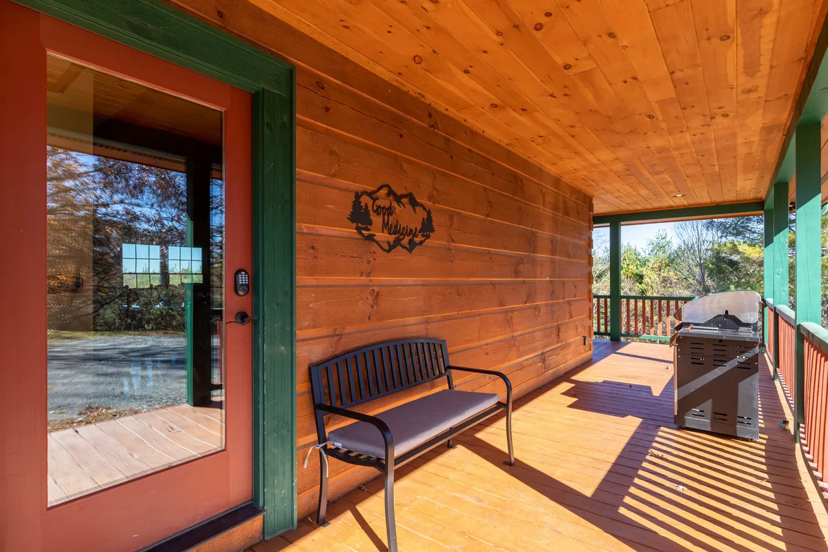 A cozy exterior porch scene featuring a wooden bench, grill station, and glass door framed by rustic wooden walls. The setup invites guests to relax outdoors, enjoy a meal, or watch sunsets from the porch, with plenty of space for social interactions.