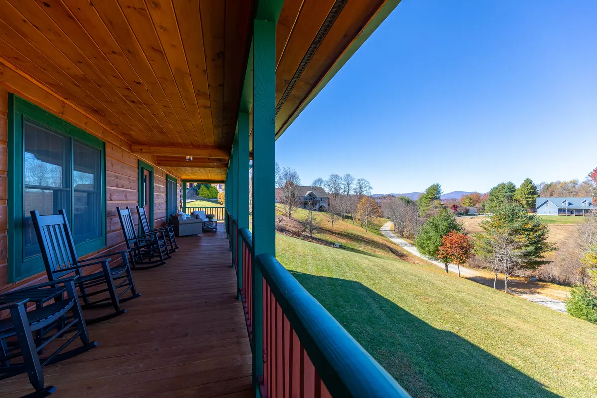 This image focuses on the inviting deck, featuring several rocking chairs lined up along the wooden railing, overlooking a lush green lawn and distant hills beneath a blue sky.