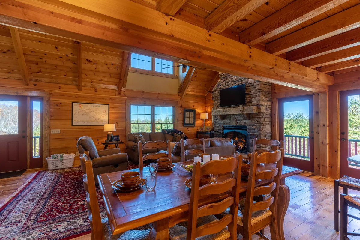 A welcoming dining area with a wooden table set for a meal, positioned near a stone fireplace, large windows that flood the space with natural light, and exposed wooden beams above. The ambiance is simultaneously elegant and homey.
