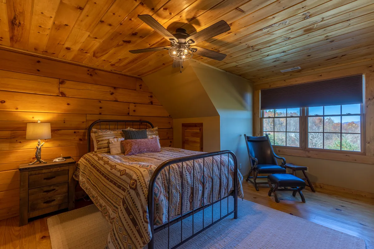 A cozy bedroom with a bed adorned in patterned bedding, a nightstand with a lamp for evening reading, a large window providing a scenic view, and an armchair in the corner. Warm and comforting, it invites relaxation.
