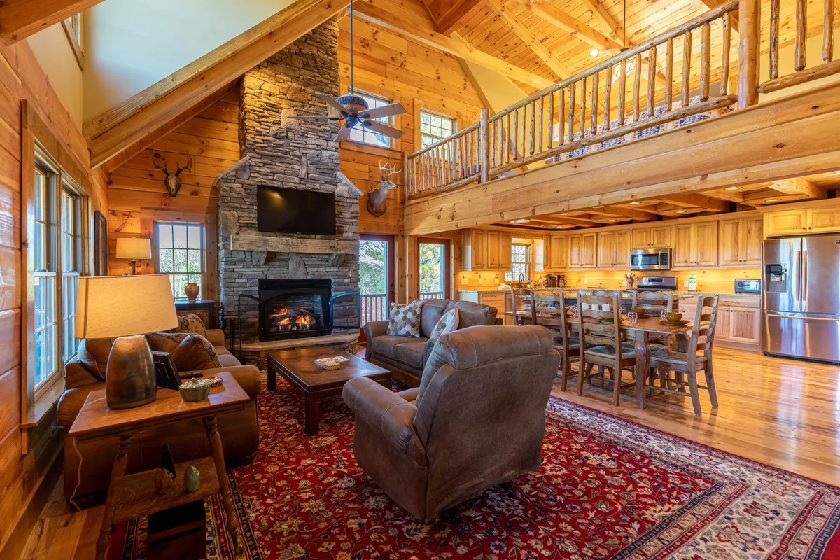 A richly detailed view of an open-concept living area featuring a dramatic stacked stone fireplace flanked by comfortable sofas, rustic wooden beams overhead, a patterned rug, and a spacious dining table adjacent to the kitchen. Warm tones and thoughtful furnishings invite relaxation and conversation.