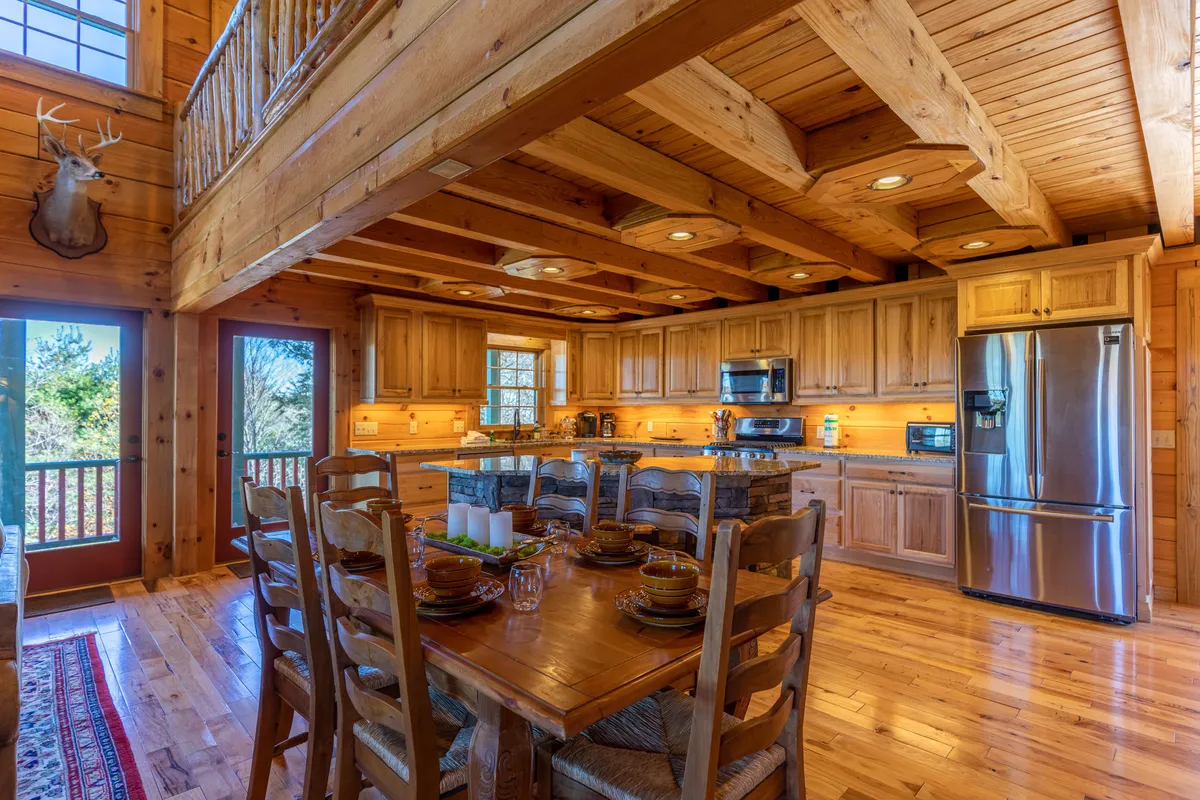 A rustic dining area with a wooden table set for a meal, adjacent to a kitchen with warm wood cabinetry and large windows offering views of the outdoors. The decor includes mountain-inspired touches like a deer mount, reinforcing the Appalachian setting.